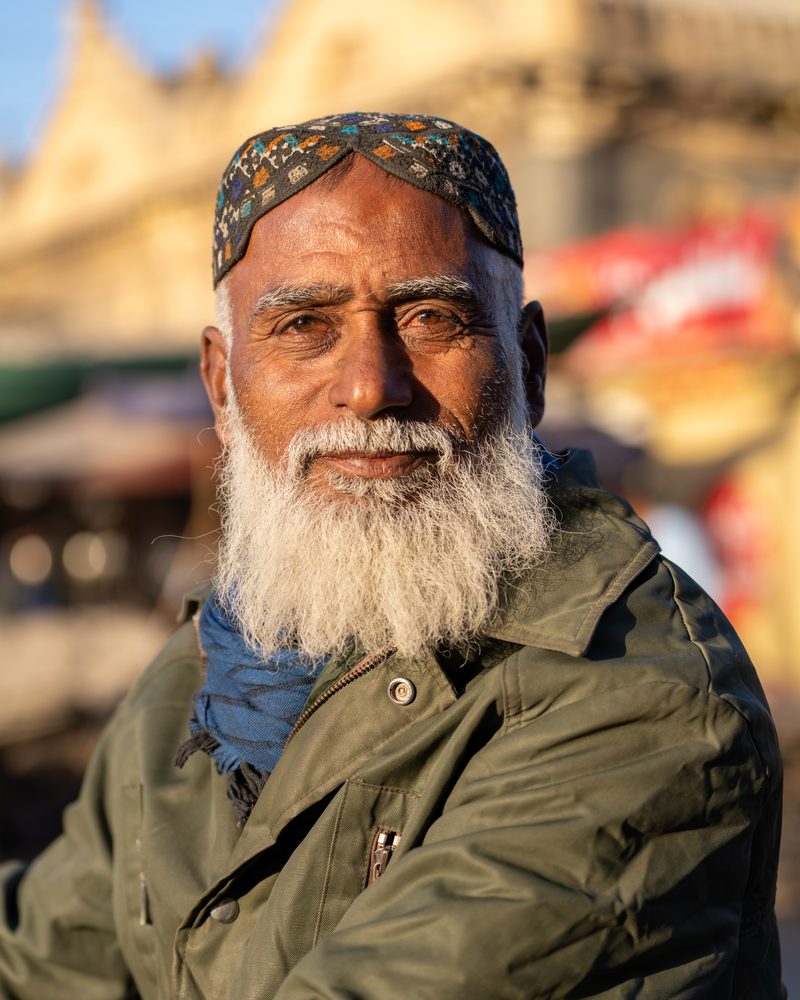 Elderly man in white kurta and cap standing with his bicycle stacked with newspapers